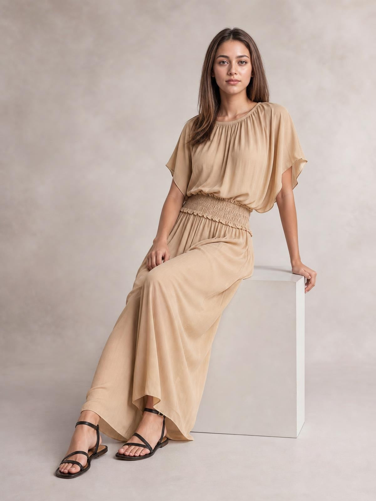 Model seated in a flowing beige chiffon maxi dress with a smocked waist, gathered neckline, flutter sleeves, and relaxed floor-length silhouette.