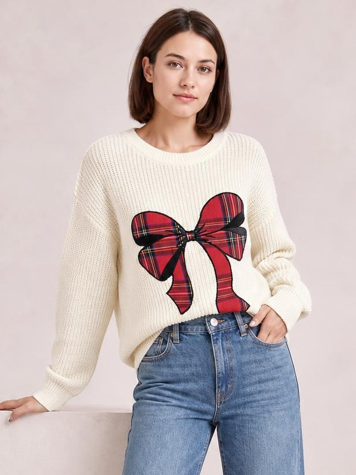 A model poses in a cream ribbed dropped-shoulder sweater with a large red plaid Christmas bow, relaxed silhouette, and ribbed cuffs and neckline.