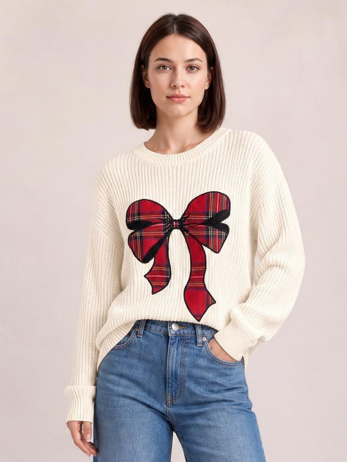 Model wears an ivory ribbed Christmas bow sweater with dropped shoulders, relaxed silhouette, crew neckline, and a large red plaid bow on the front.