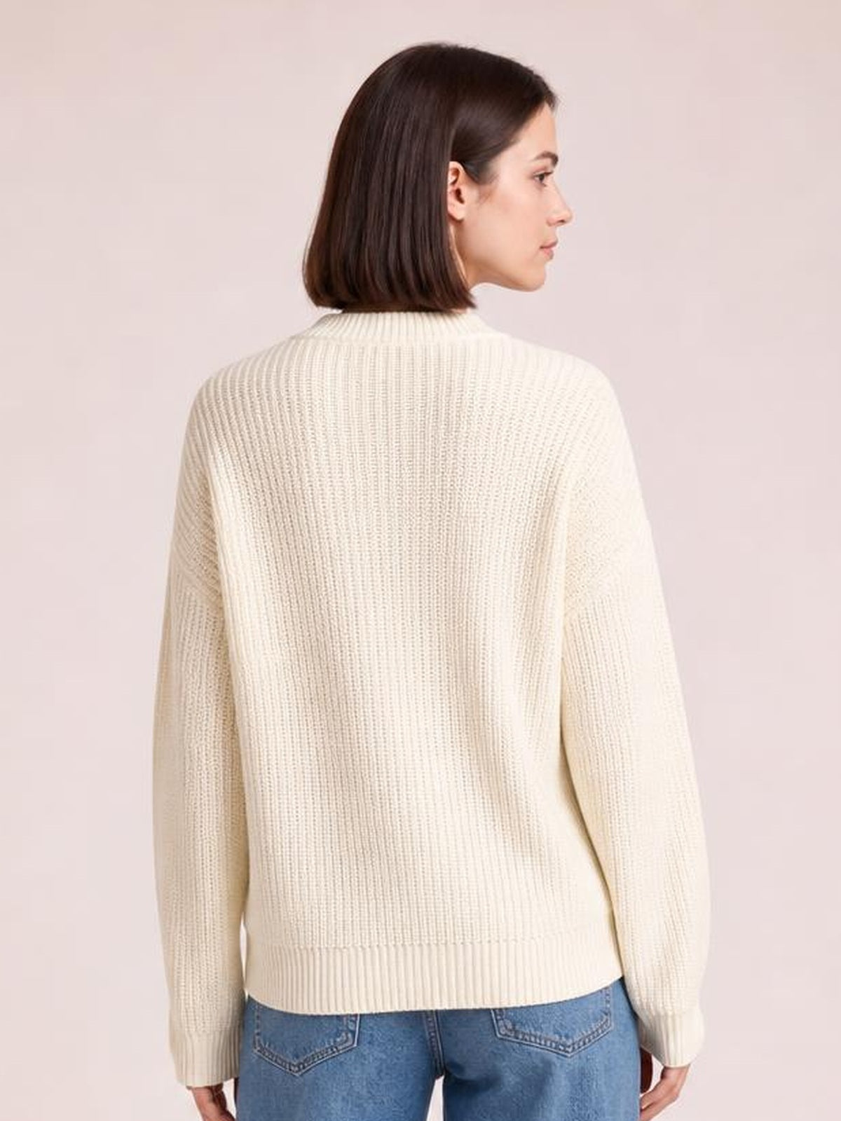 A model poses in a cream ribbed sweater with dropped shoulders, long sleeves, a high neckline, relaxed silhouette, and a wide ribbed hem.