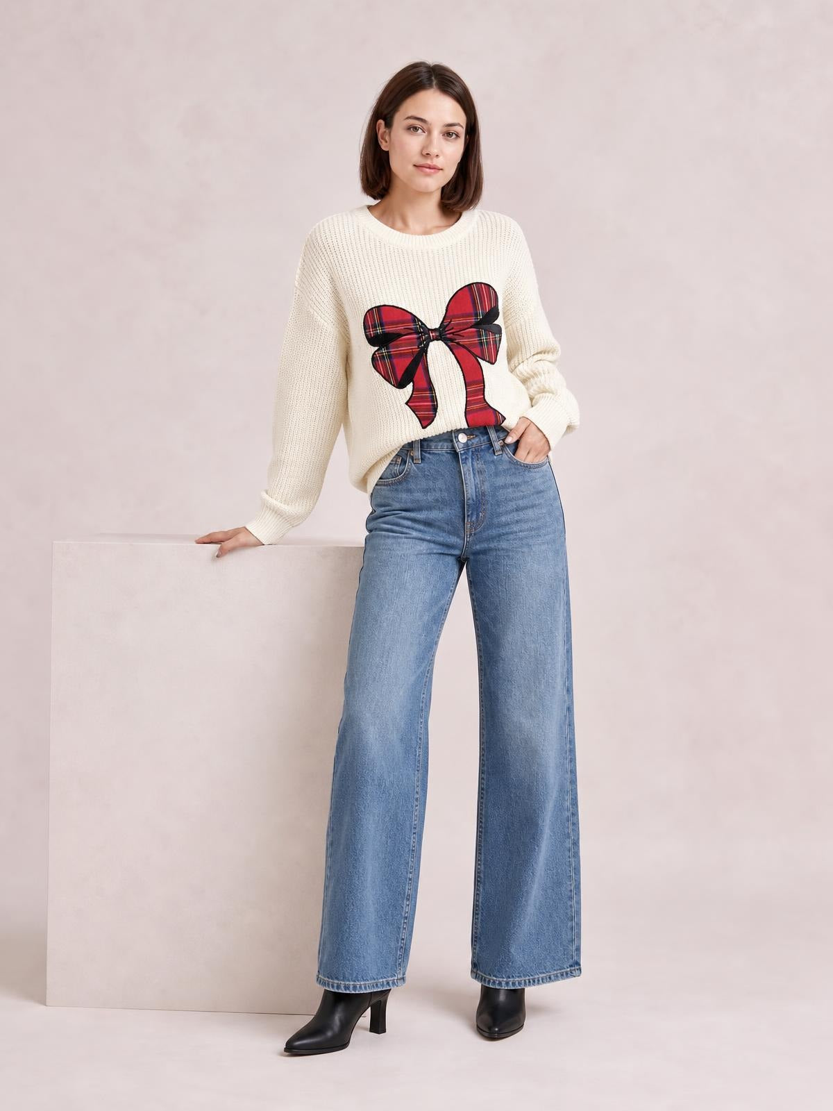 A model poses in a cream ribbed dropped-shoulder sweater with a large red plaid Christmas bow, relaxed fit, and crew neckline.