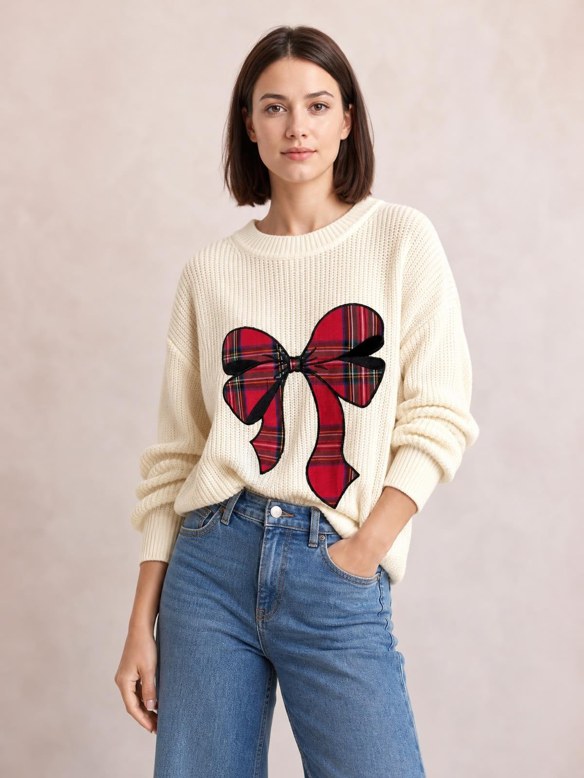 Model wearing an ivory ribbed dropped-shoulder sweater with a large red plaid Christmas bow, relaxed silhouette, and rolled cuffs while posing with one hand in her jeans pocket.