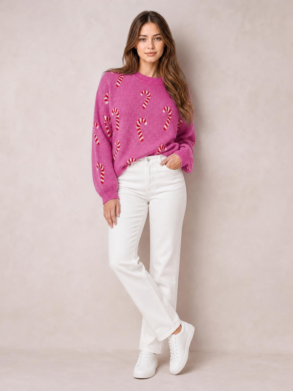 A model poses in a pink Christmas drop-shoulder sweater with candy-cane motifs, ribbed crew neck and cuffs, relaxed silhouette, and one hand in her pocket.