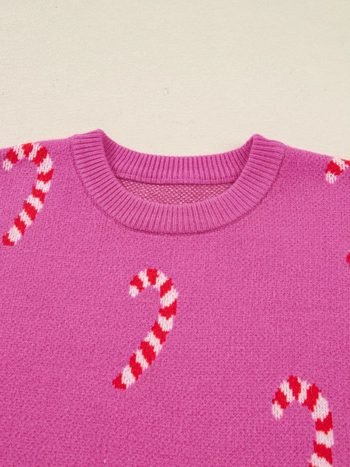 Close-up of a pink knit holiday sweater with a ribbed crew neckline and red-and-white candy cane motifs across the drop-shoulder design.