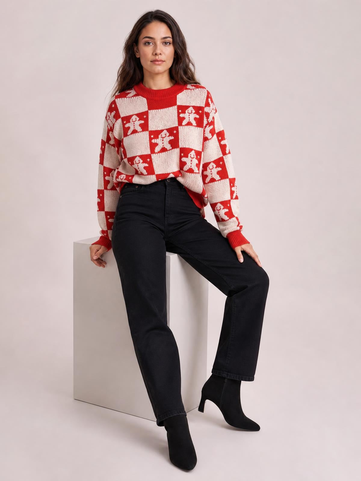 A model sits wearing a red-and-cream checkerboard Christmas sweater with gingerbread figures, round neck, long sleeves, and ribbed cuffs.