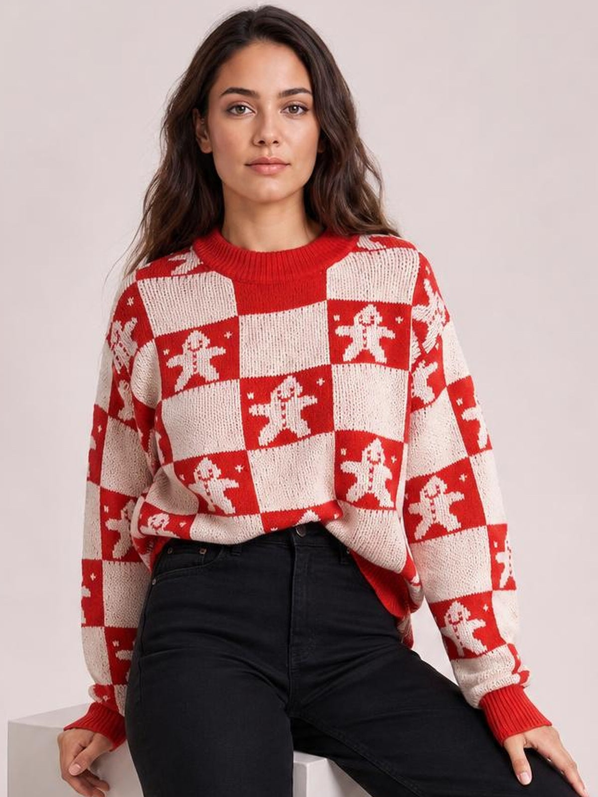 A seated model wears a red and cream Christmas checkerboard sweater with gingerbread motifs, round neck, long sleeves, ribbed cuffs, and a relaxed silhouette.