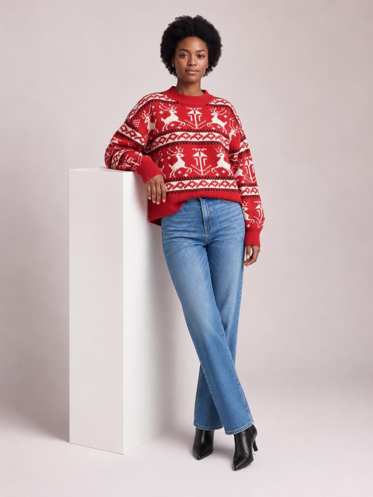 Model leans against a pedestal wearing a red Christmas sweater with a round neck, long sleeves, reindeer pattern, relaxed fit, and ribbed cuffs and hem.