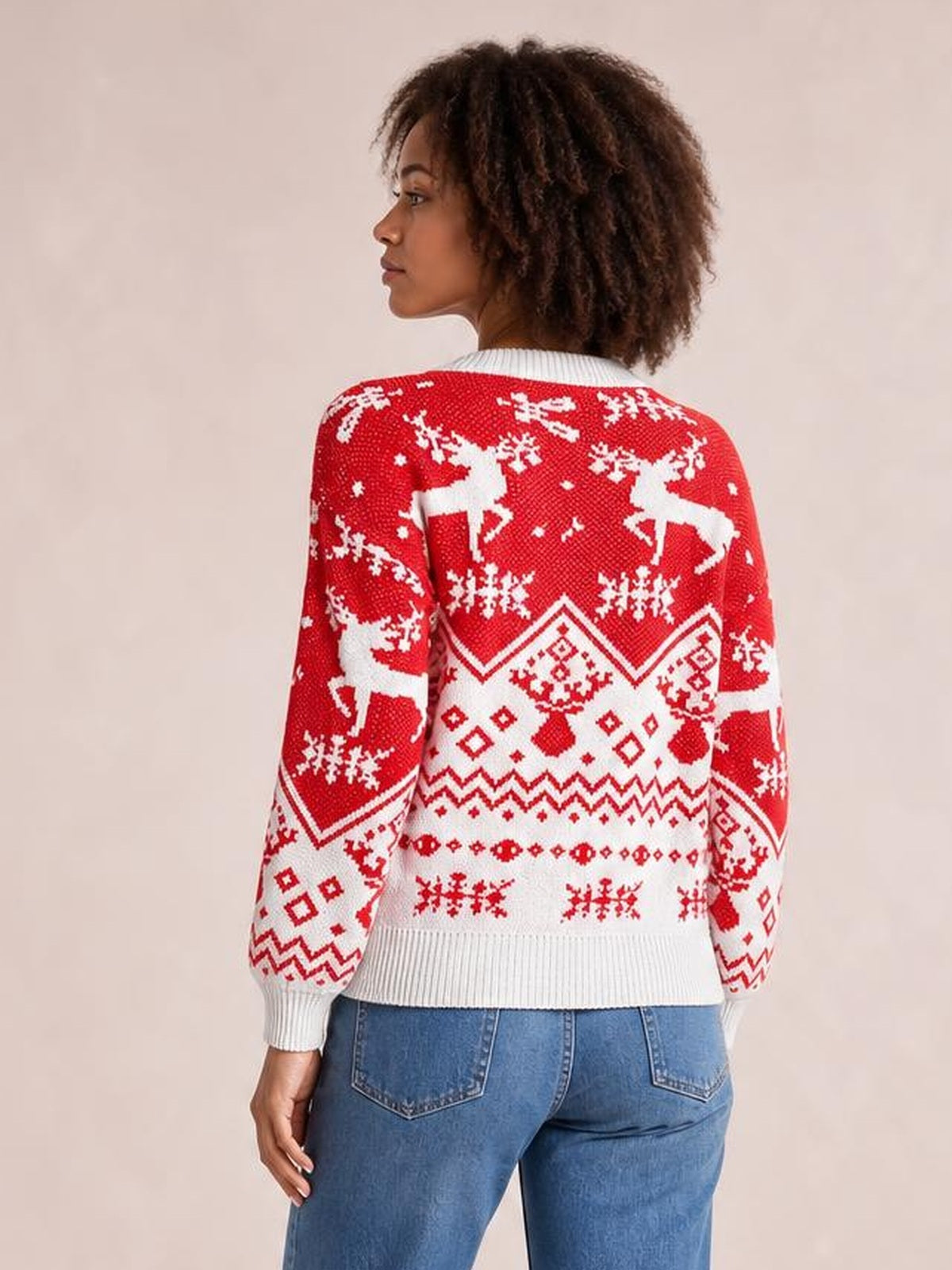 Model turned to show the back of a red Christmas sweater with white reindeer, snowflake and geometric motifs, long sleeves, a round neck, and ribbed cuffs and hem.