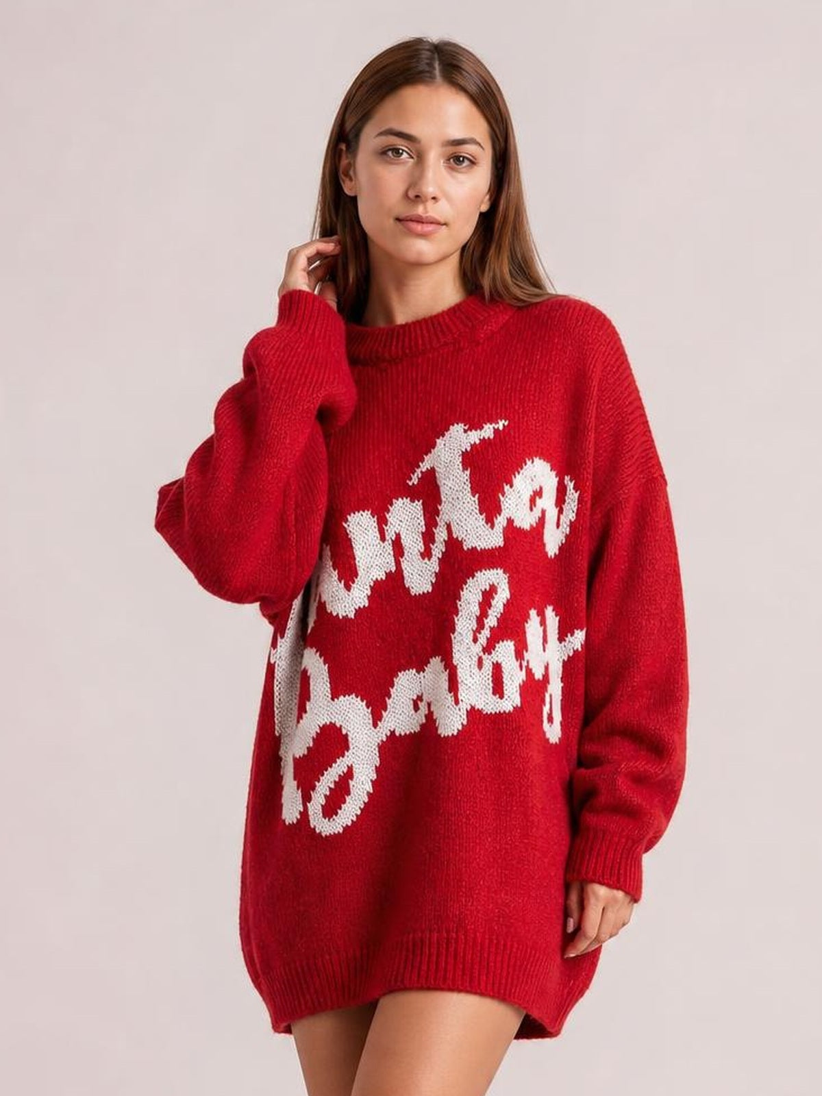 Model poses in a red oversized Christmas letter sweater dress with white festive lettering, ribbed crew neckline, cuffs, and hem.