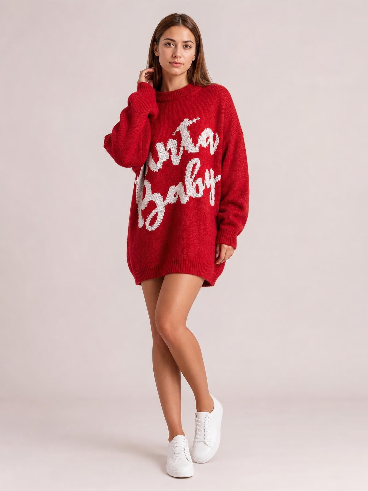 A model poses in a red oversized sweater dress with white Christmas lettering, crew neckline, long sleeves, ribbed cuffs and hem, and a relaxed silhouette.