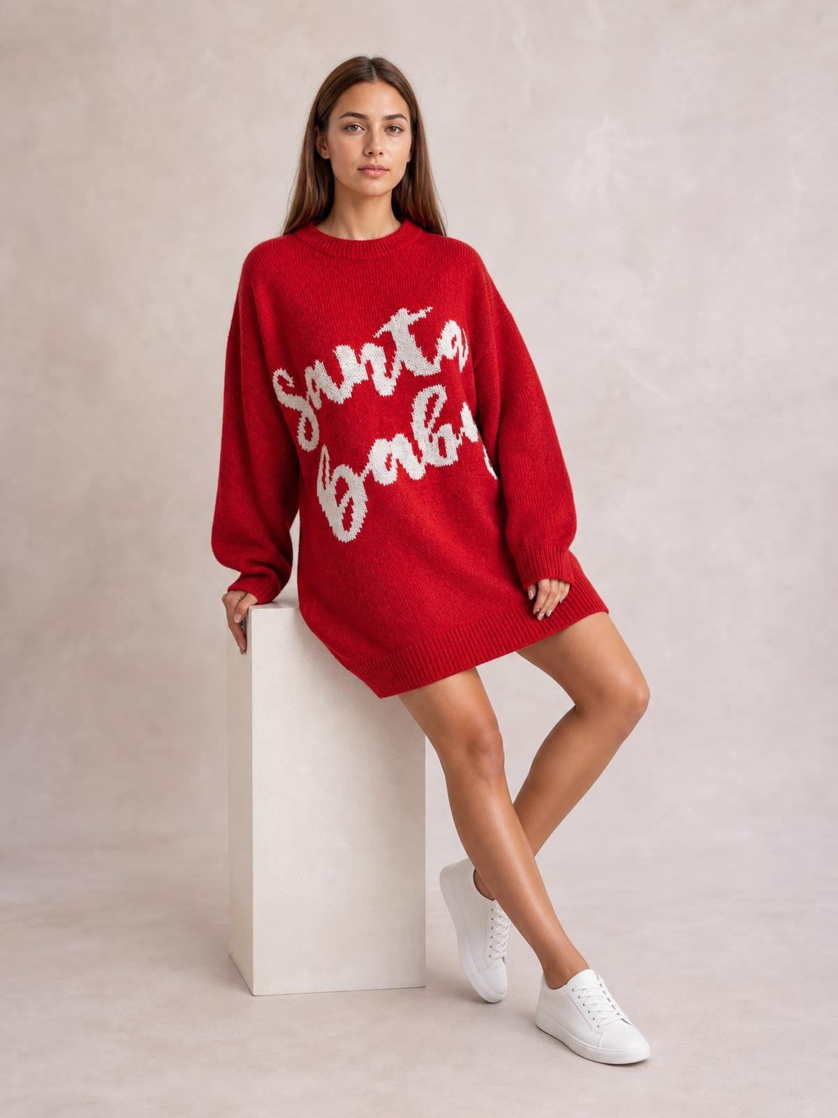 A model sits in a red oversized Christmas sweater dress with white festive lettering, a crew neckline, long sleeves, and ribbed cuffs and hem.