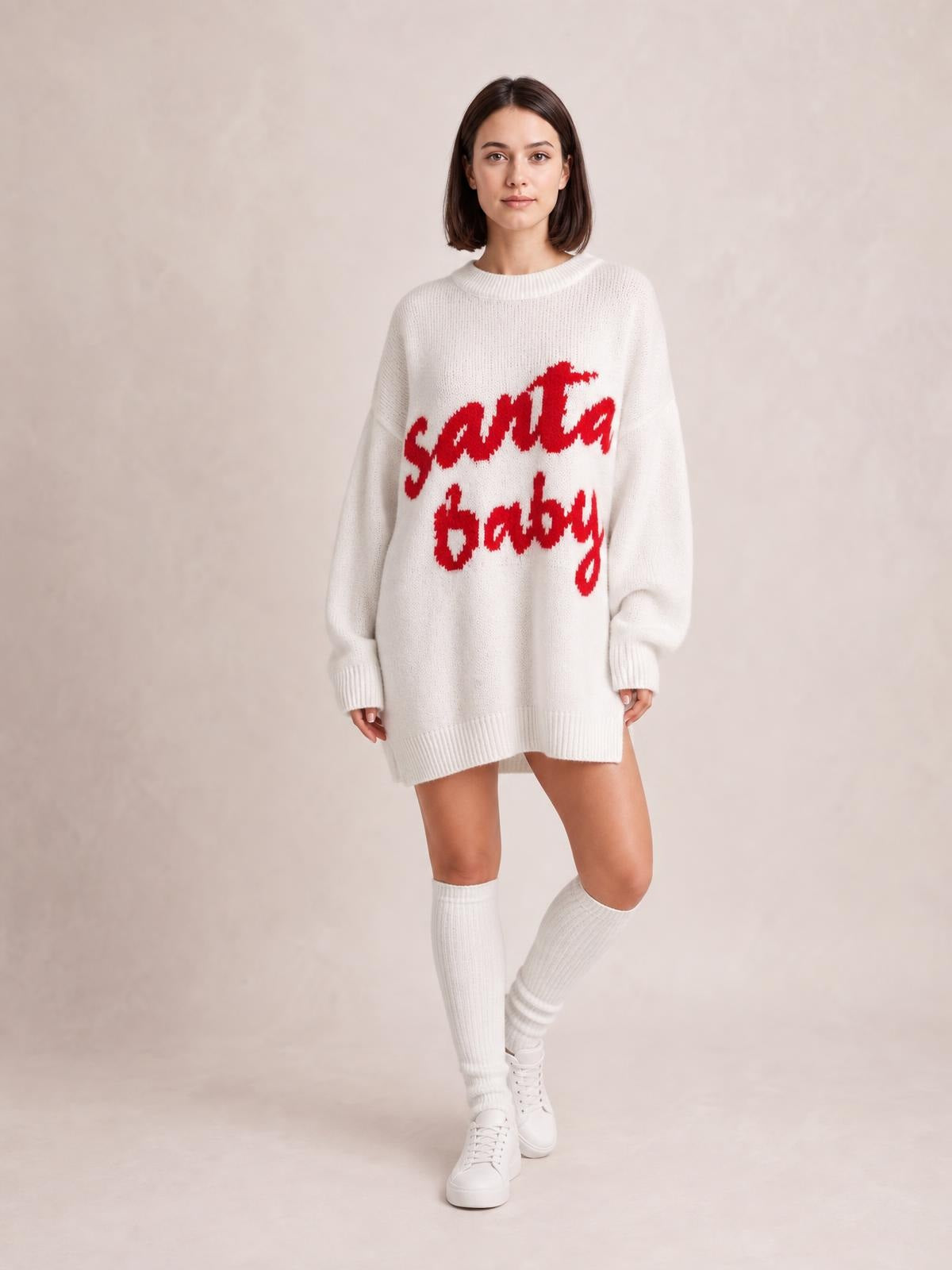 A model poses in a white oversized Christmas sweater dress with red “Santa Baby” lettering, crew neckline, long sleeves, and ribbed cuffs and hem.