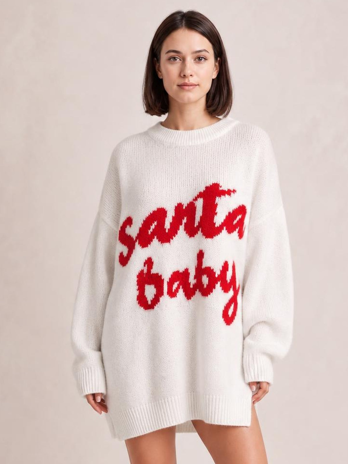 Model stands facing forward in an oversized white Christmas sweater dress with red “Santa Baby” lettering, long sleeves, ribbed crew neck, cuffs, and hem.