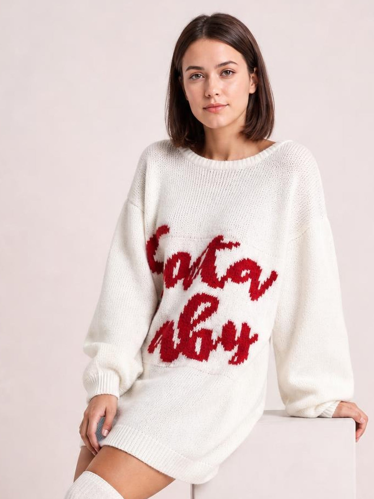 A model sits in a white oversized Christmas sweater dress with bold red lettering, a round neckline, long sleeves, and ribbed cuffs and hem.