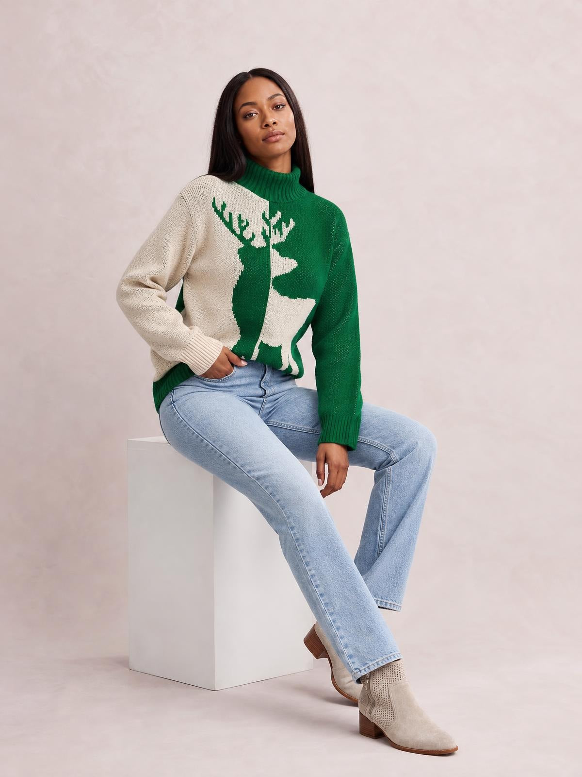 Model seated in light jeans wears a ribbed green and cream Christmas reindeer turtleneck sweater with long sleeves and a relaxed silhouette.