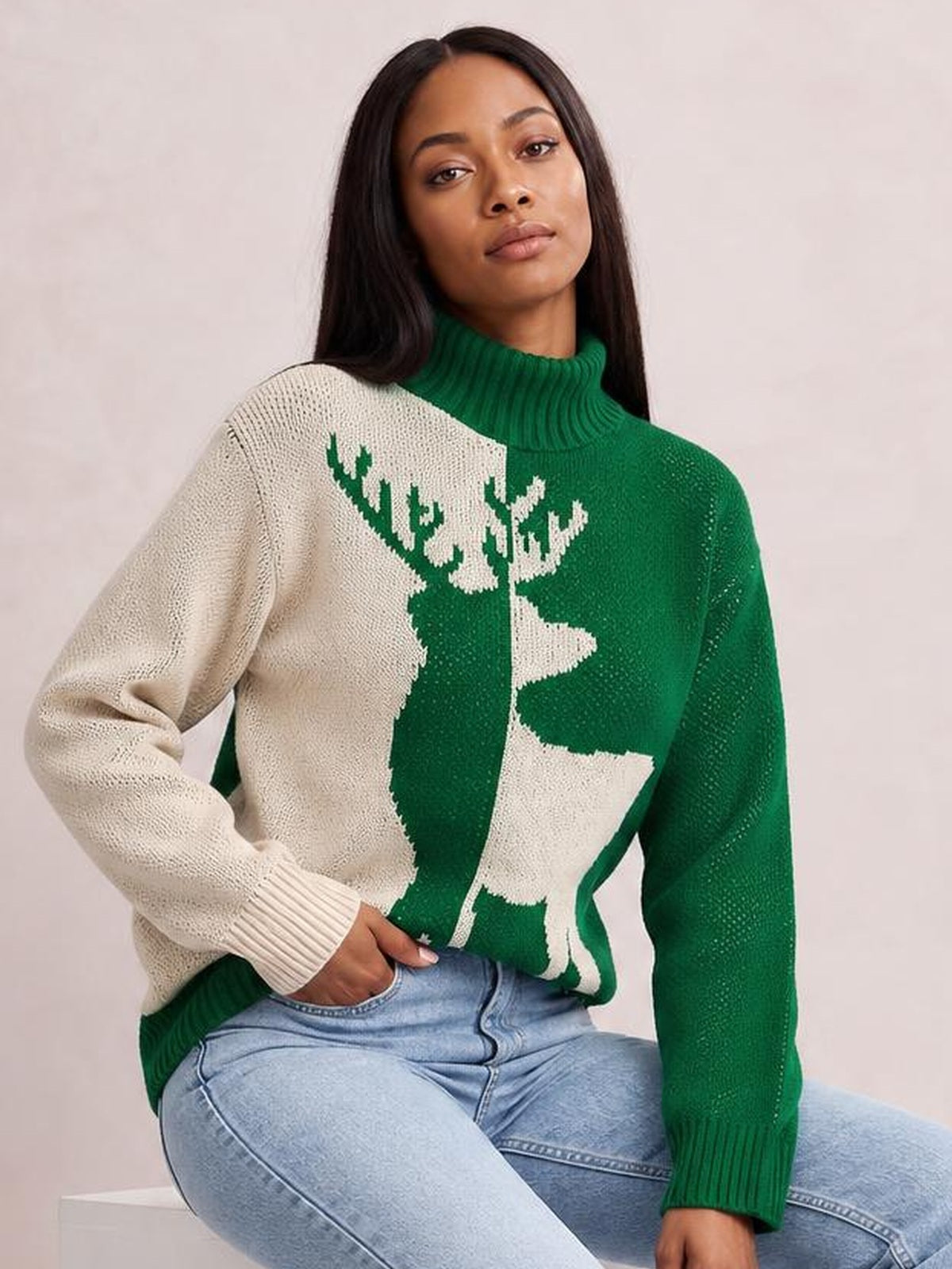 Model seated in light-wash jeans wears a green and cream ribbed turtleneck sweater with a large reindeer intarsia and long sleeves.
