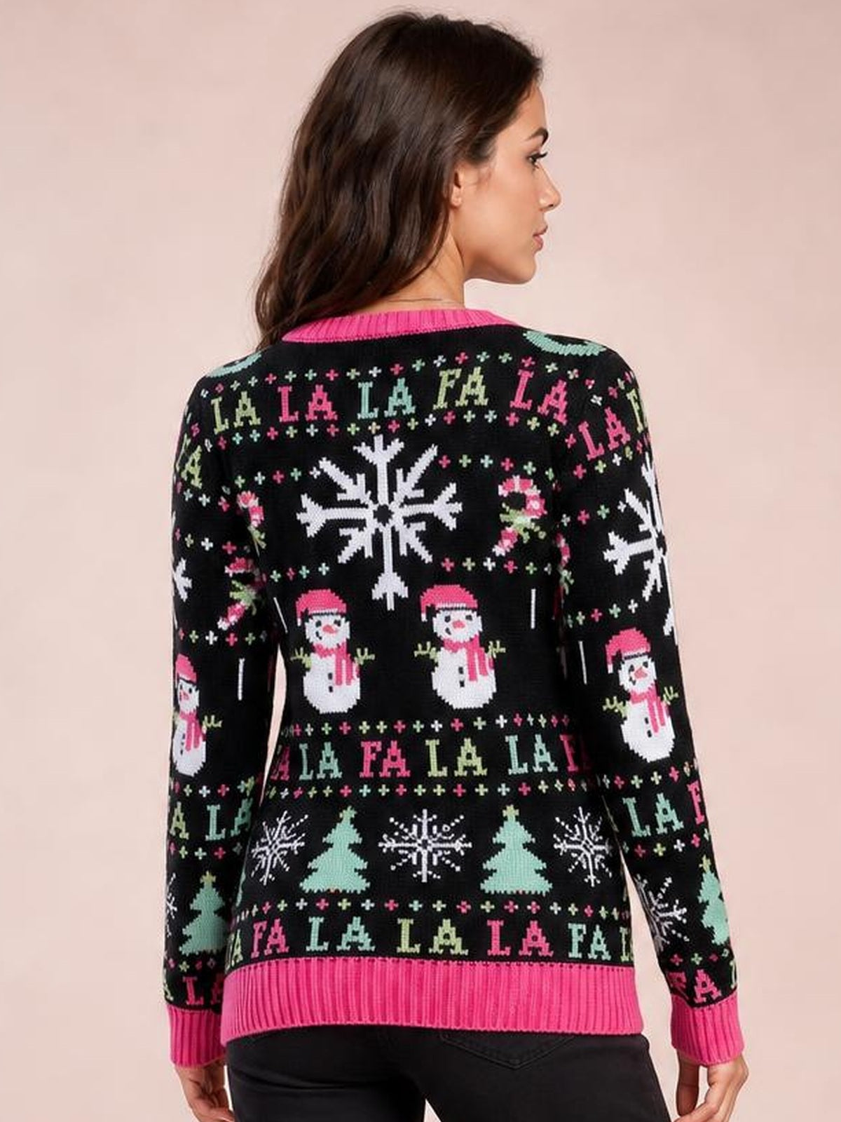 A woman models the back of a black Christmas sweater with pink ribbed trim, long sleeves, and colorful snowflake, snowman, tree, and “FA LA LA” motifs.