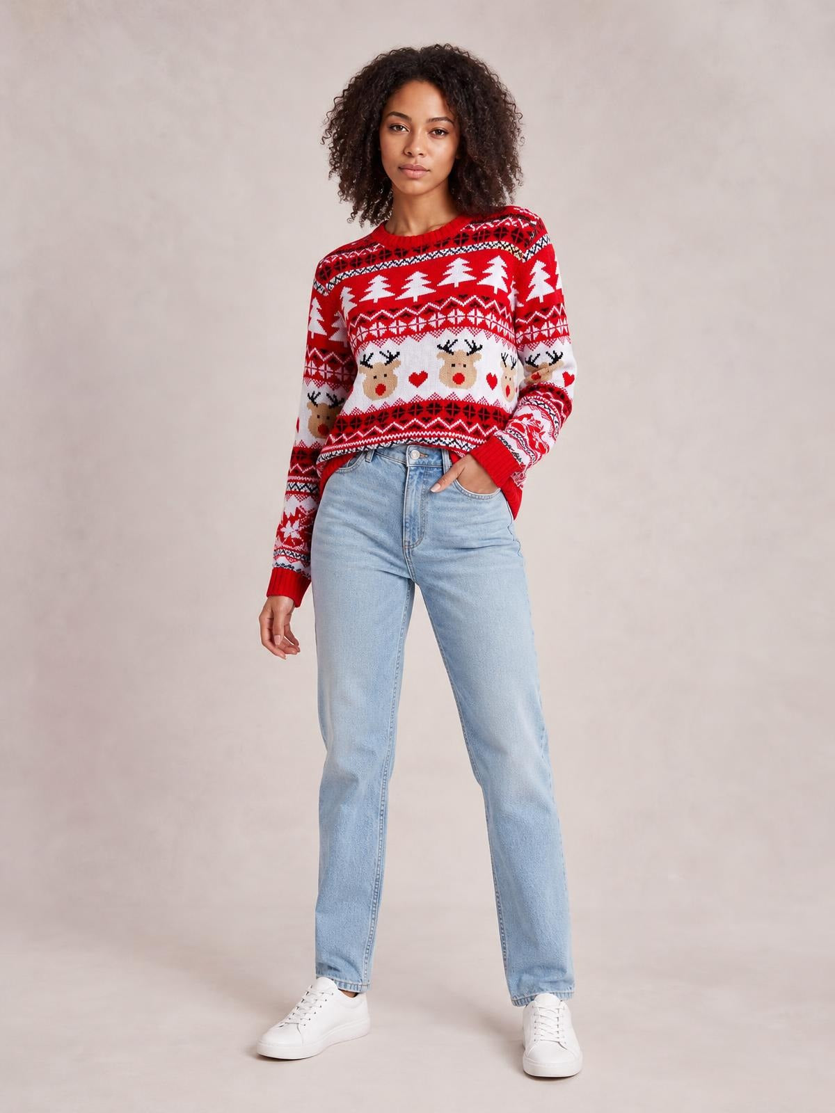 A model stands in light-wash jeans wearing a red and white Christmas round neck sweater with reindeer, tree, and geometric motifs, long sleeves, and ribbed cuffs.