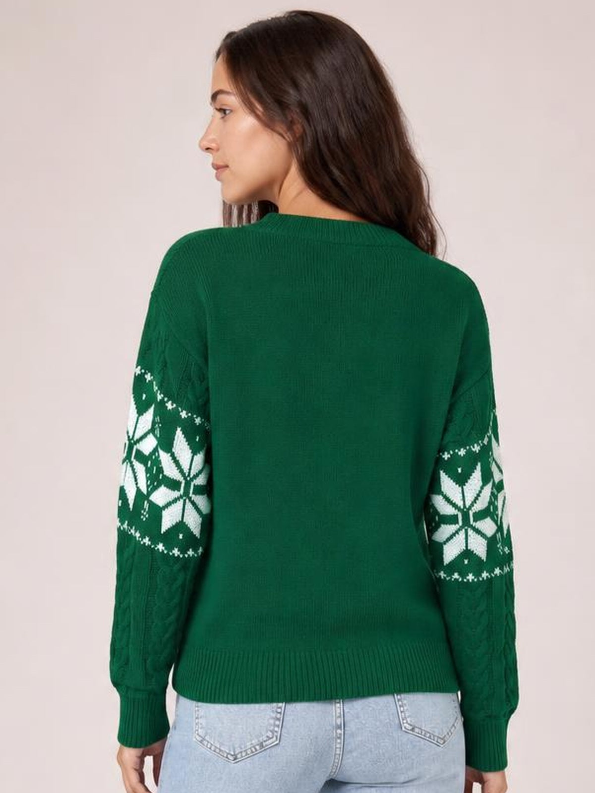 Model shown from behind in a relaxed green cable-knit sweater with white snowflake-patterned sleeves, a round neckline, ribbed cuffs, and hem.