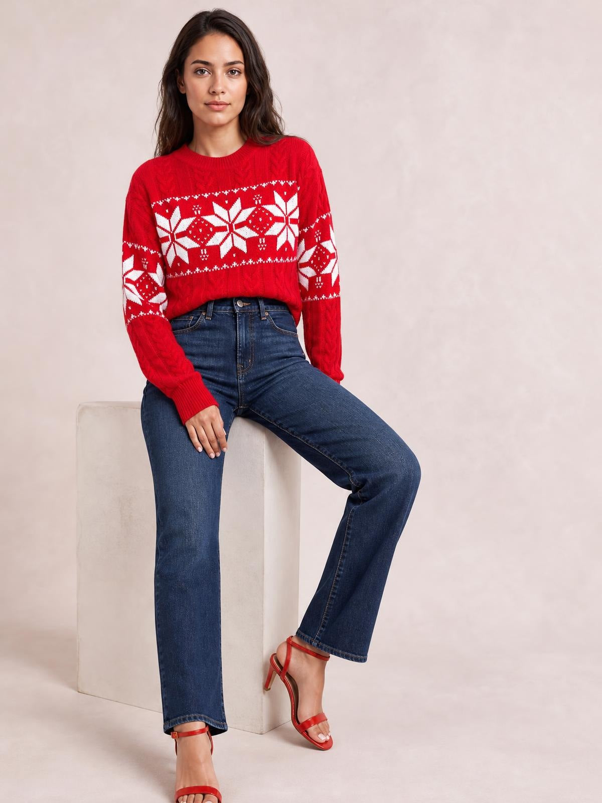 A woman poses seated in a bright red cable-knit Christmas sweater with white snowflake patterns, ribbed crew neck and cuffs, and a relaxed silhouette.