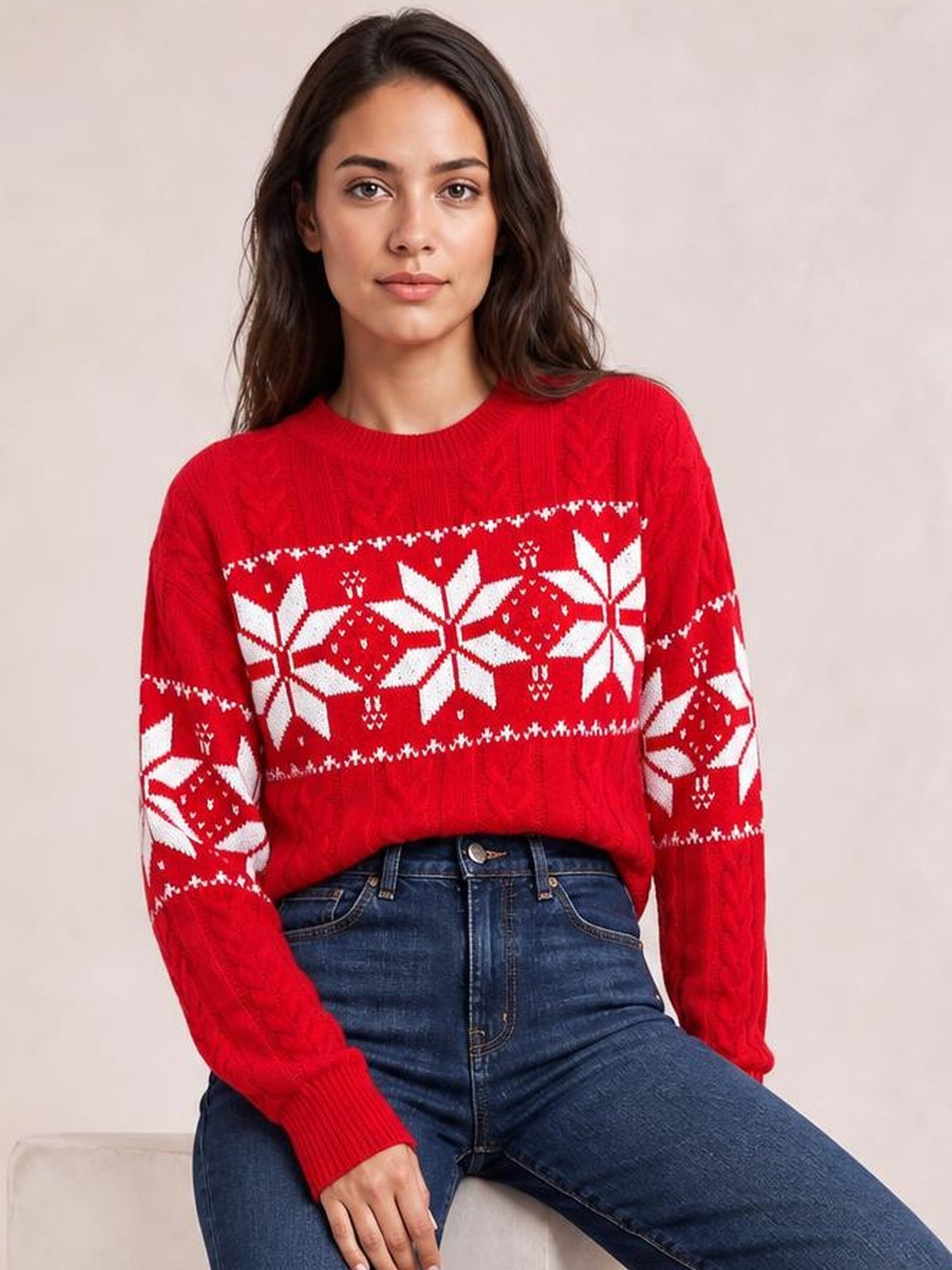 Model seated in a red cable-knit Christmas sweater with white snowflake patterns, crew neckline, long sleeves, ribbed cuffs, and a relaxed silhouette.