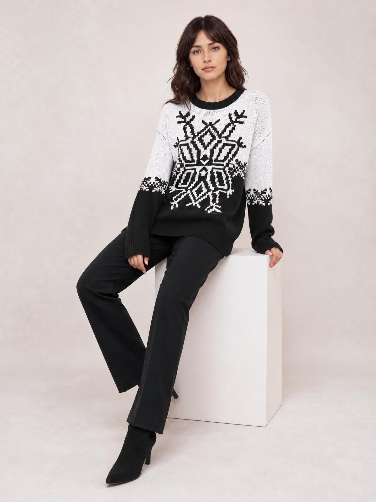 Woman seated in a relaxed pose wearing a black-and-white Christmas snowflake round-neck sweater with a bold geometric motif, ribbed cuffs, and hem.