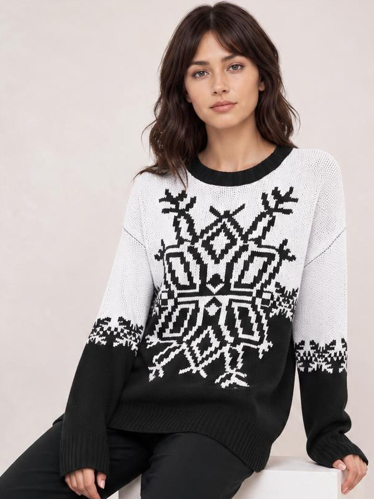 A seated model wears a black-and-white Christmas snowflake round-neck sweater with a relaxed silhouette, ribbed cuffs and hem, and long sleeves.