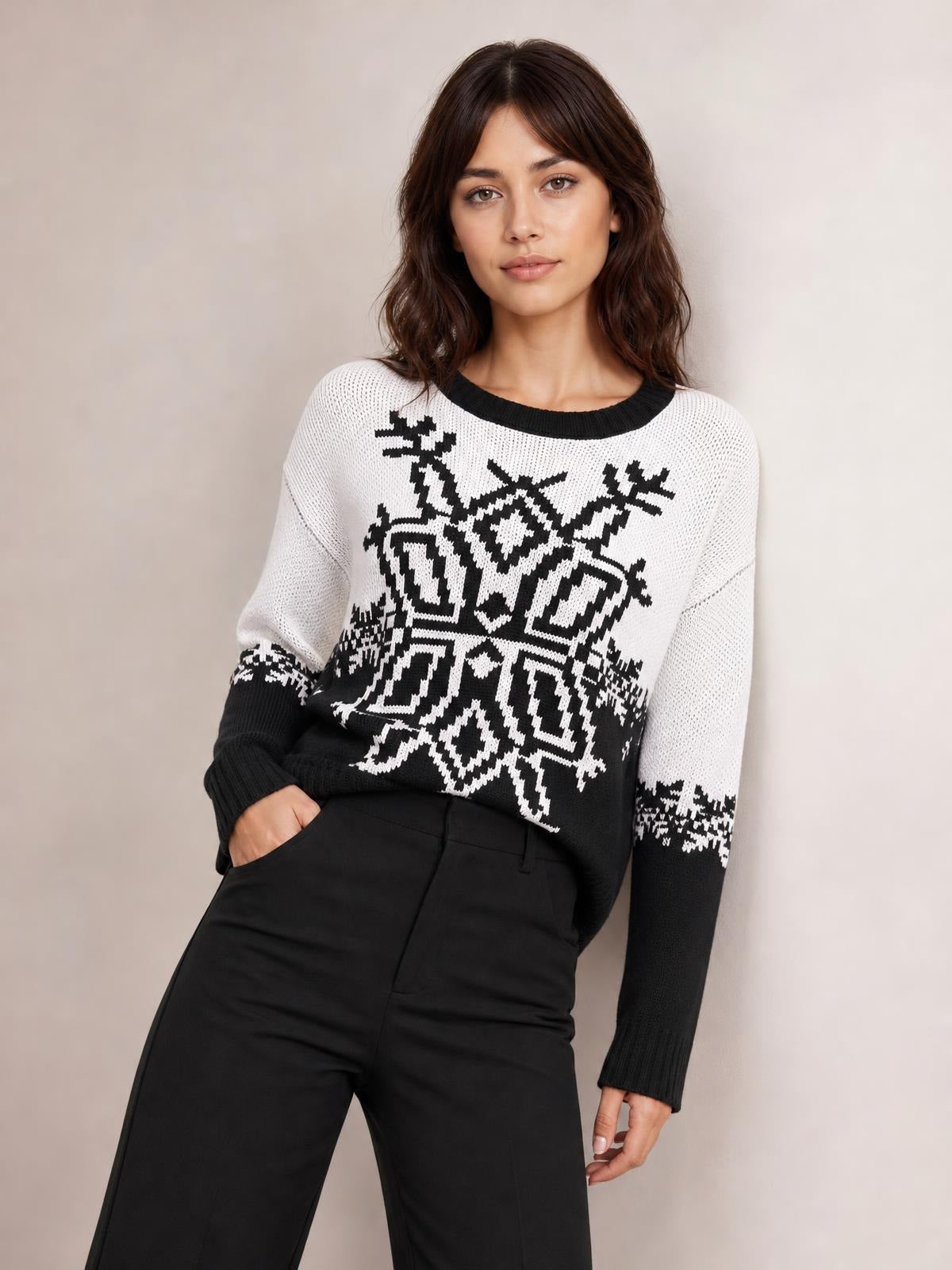 Model wears a white and black Christmas snowflake round neck sweater with long sleeves, ribbed cuffs, and a relaxed silhouette, posing with one hand in her pocket.
