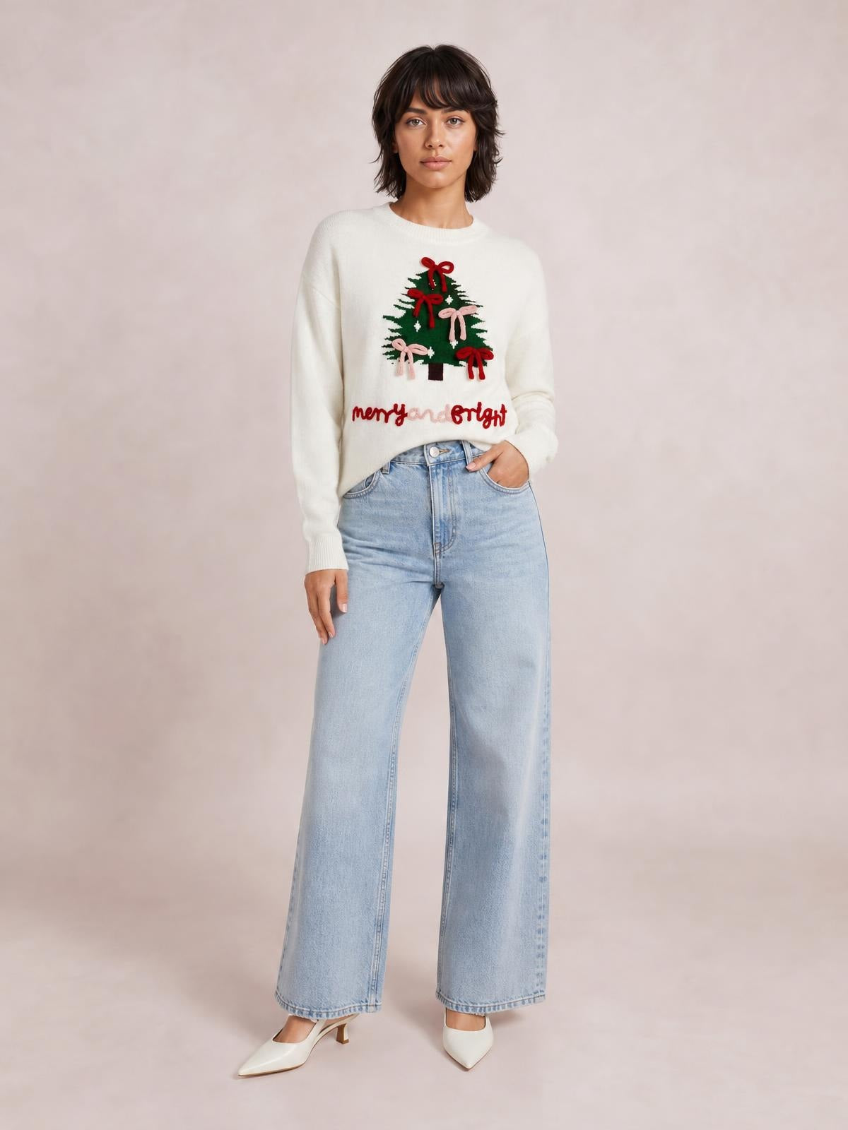Model poses in a cream drop-shoulder knit sweater featuring a Christmas tree decorated with colorful bows and “merry and bright” lettering.