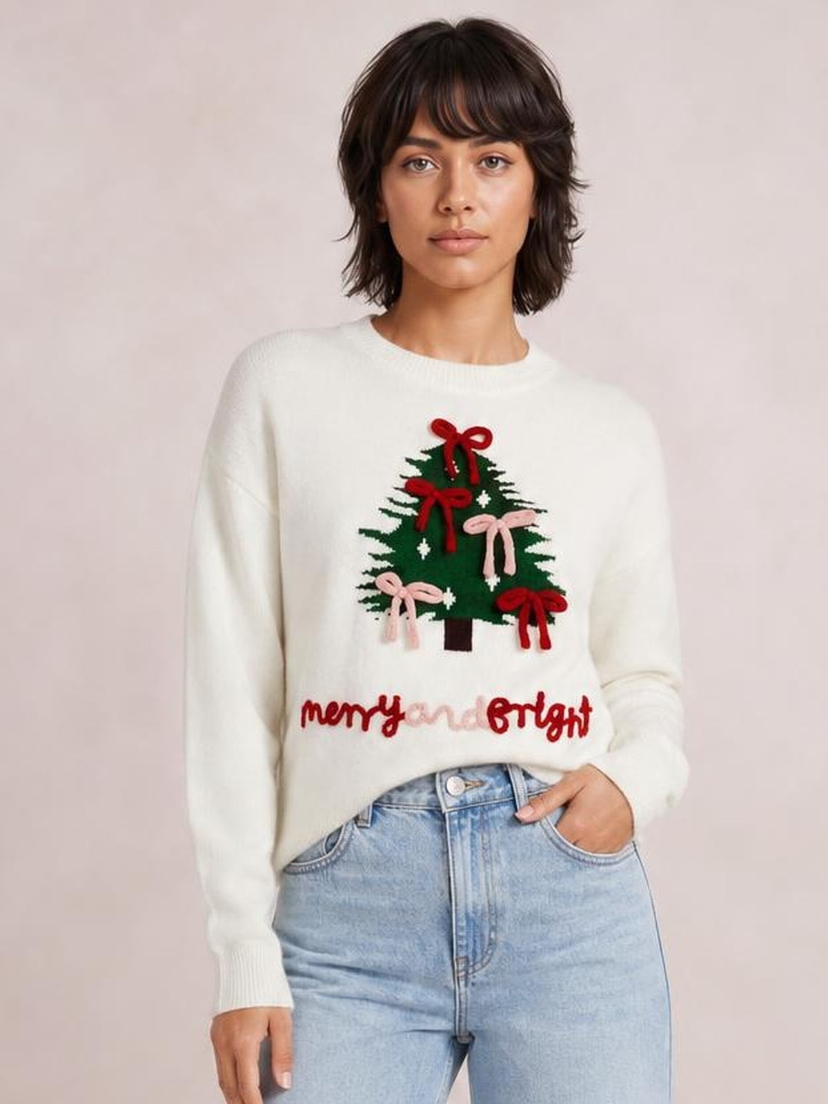 Model wearing an ivory drop-shoulder knit sweater with a Christmas tree and red and pink bow design, ribbed crew neck, cuffs, and hem.