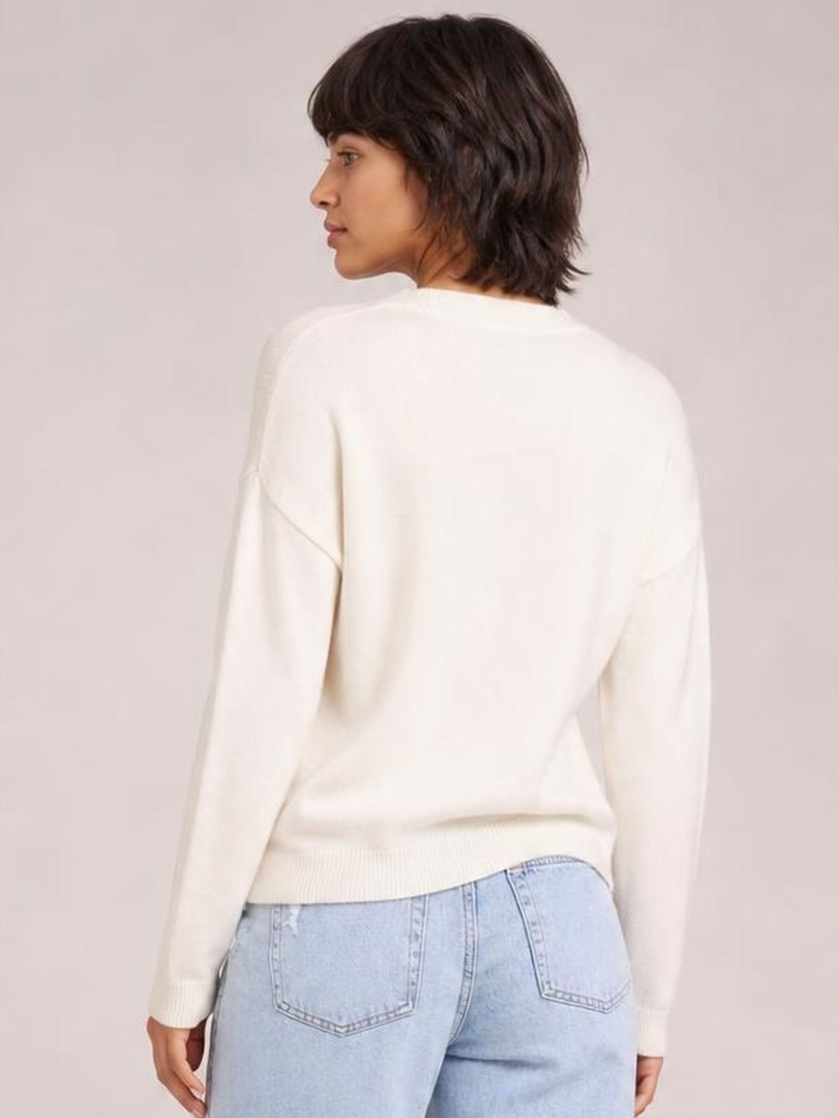 Model turns in profile to show the back of a cream drop-shoulder knit sweater with long sleeves, ribbed cuffs and a ribbed hem.