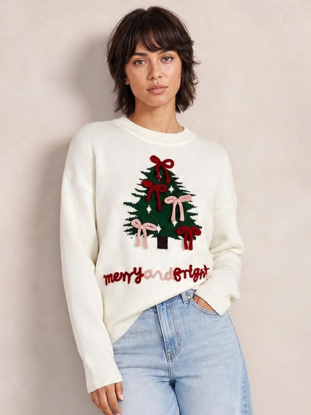 A model wears a white drop-shoulder knit sweater with a relaxed silhouette, ribbed trims, and a Christmas tree graphic decorated with red and pink bows.