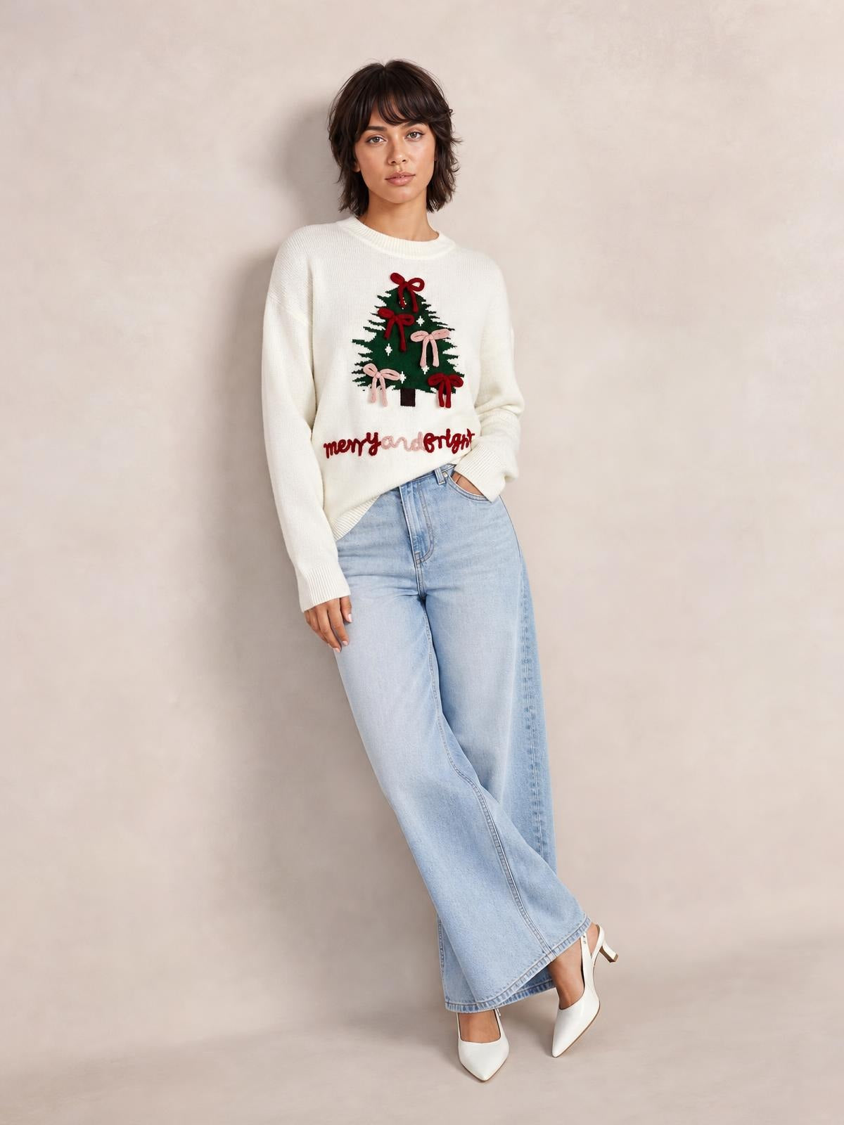A model wears a cream Christmas tree bow drop-shoulder knit sweater with a crew neck, ribbed cuffs, festive tree appliqué, and a relaxed silhouette.