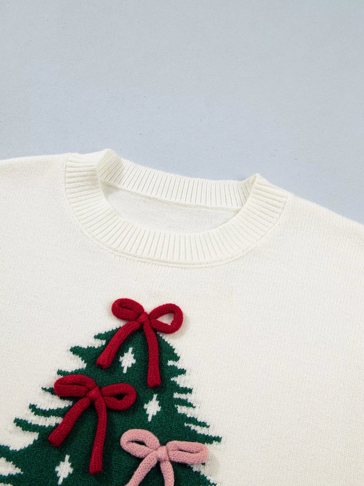 Close-up of an ivory knit sweater with a ribbed crew neckline and a green Christmas tree applique decorated with red and pink bow details.