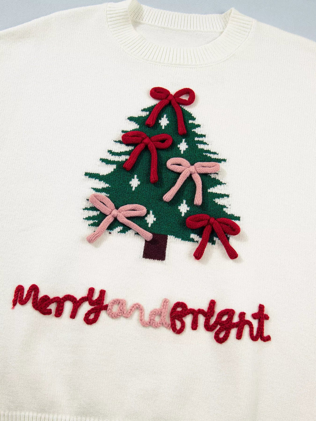 Close-up of a white knit sweater with a ribbed crew neck and a colorful Christmas tree applique decorated with red and pink bows.