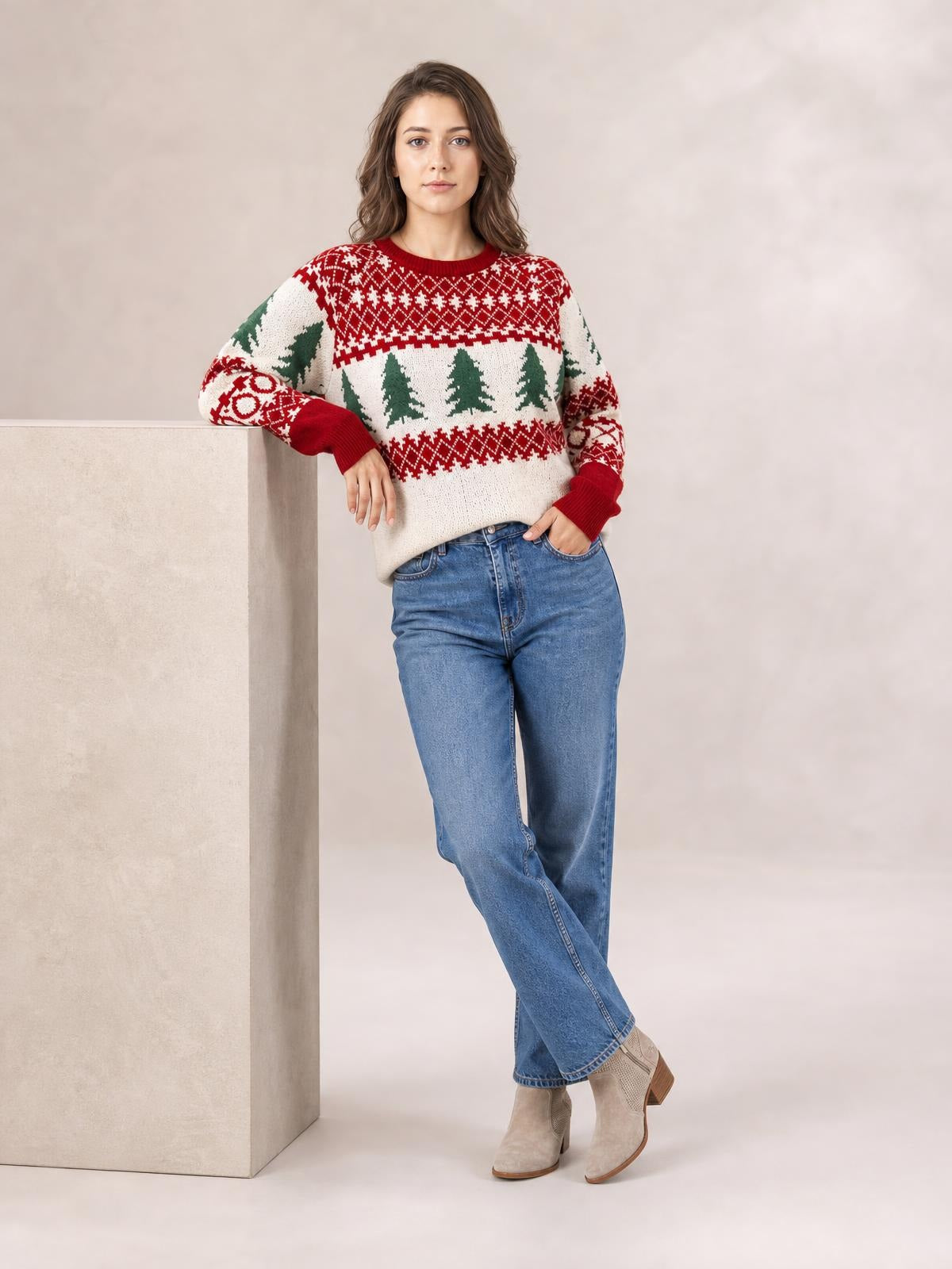 Model leans against a pedestal wearing a cream Christmas tree pattern round neck sweater with red and green motifs, ribbed cuffs, and a relaxed silhouette.