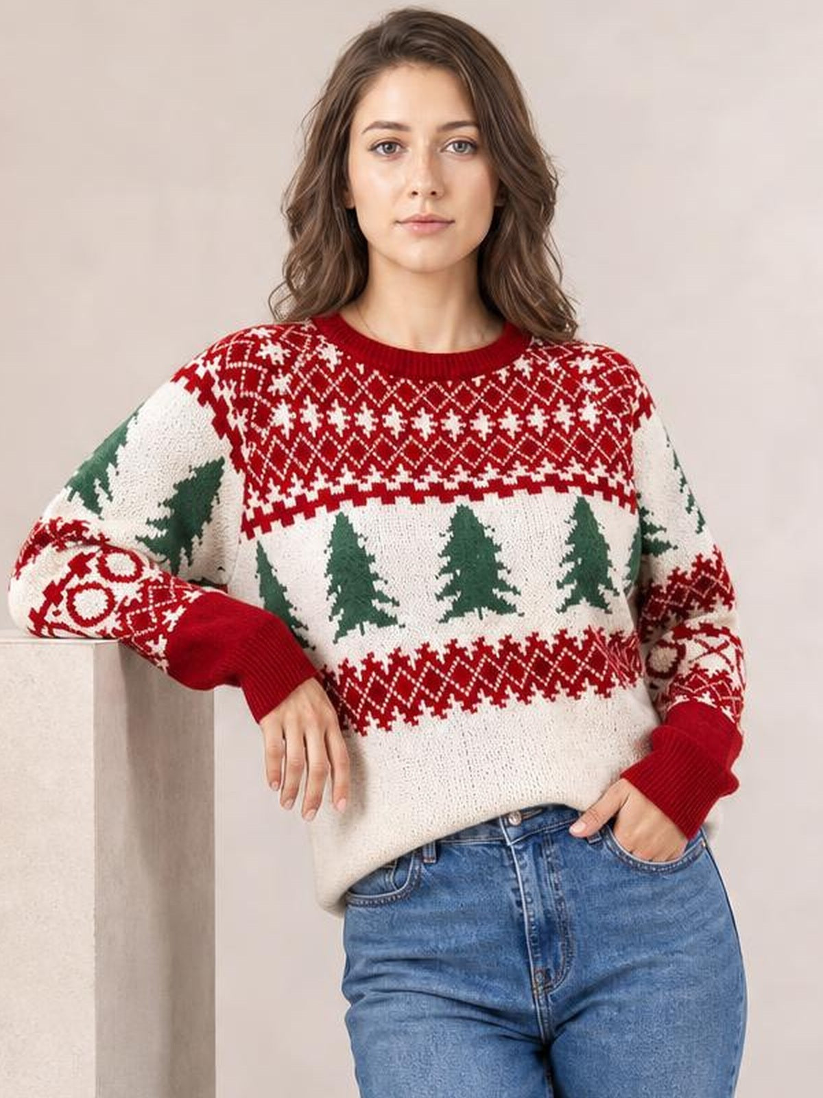 A model wears a relaxed Christmas tree pattern round neck sweater in cream, red, and green with ribbed cuffs and hem, posed with one hand in her jeans pocket.