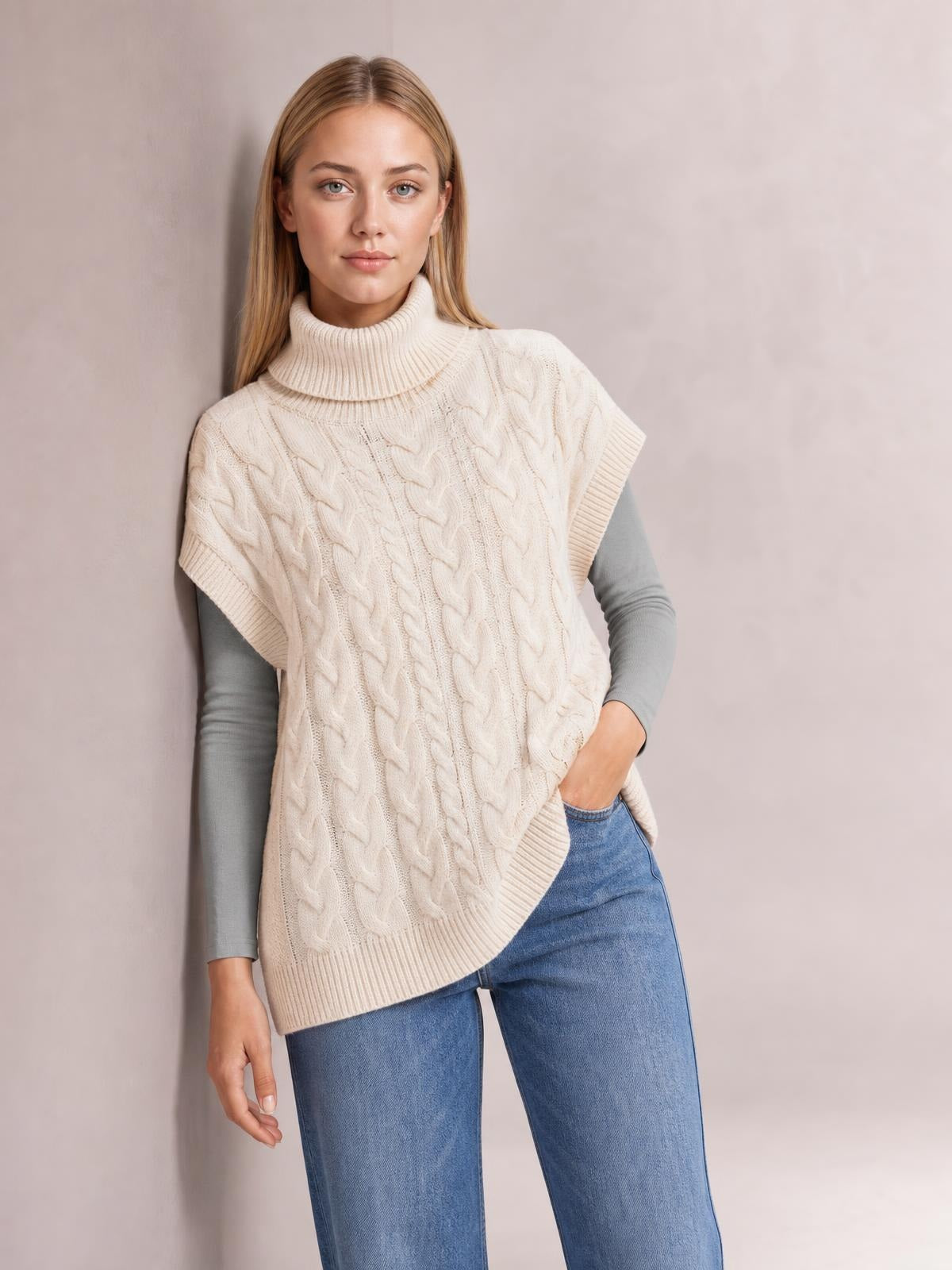Model wearing an ivory chunky knit turtleneck cap sleeve sweater with cable-knit texture, ribbed trim, and a relaxed tunic silhouette.