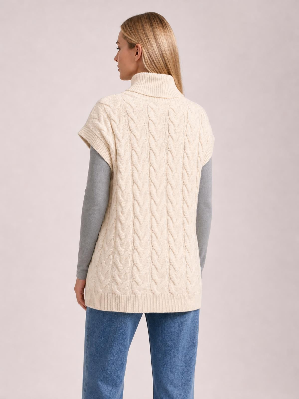 A model shown from behind wears a cream chunky cable-knit turtleneck cap-sleeve sweater with a relaxed, tunic-length silhouette and ribbed hem.