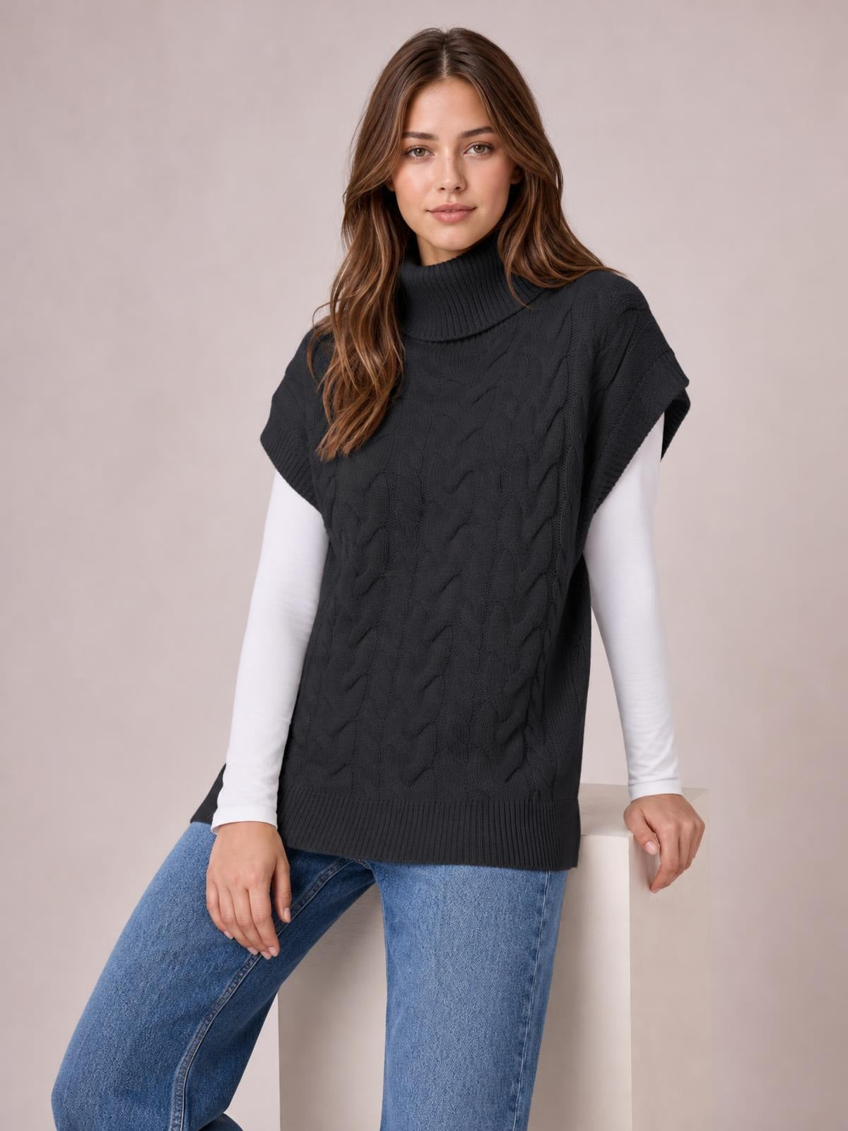 A model wears a black chunky cable-knit turtleneck sweater with cap sleeves, ribbed trim, and a relaxed long silhouette over a white top and jeans.