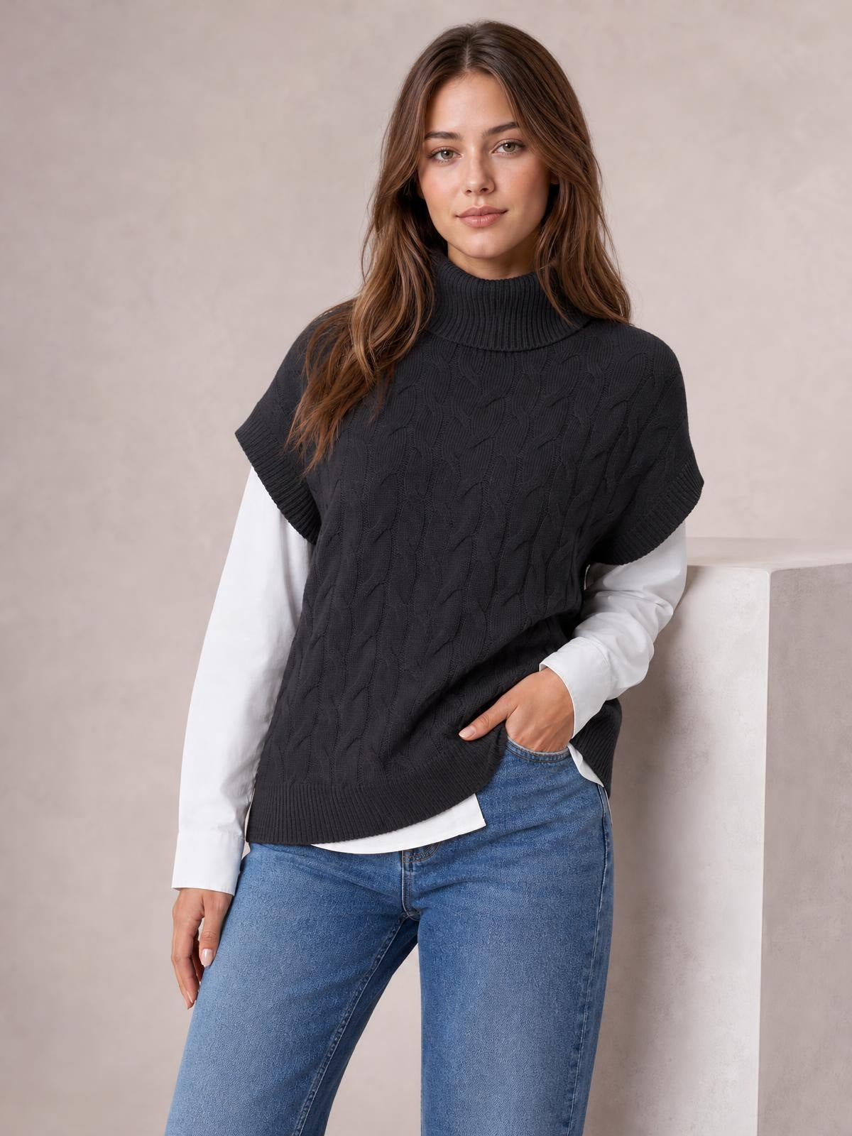 Model wears a black chunky cable-knit turtleneck sweater with cap sleeves, ribbed trim, and a relaxed silhouette over a white shirt and jeans.