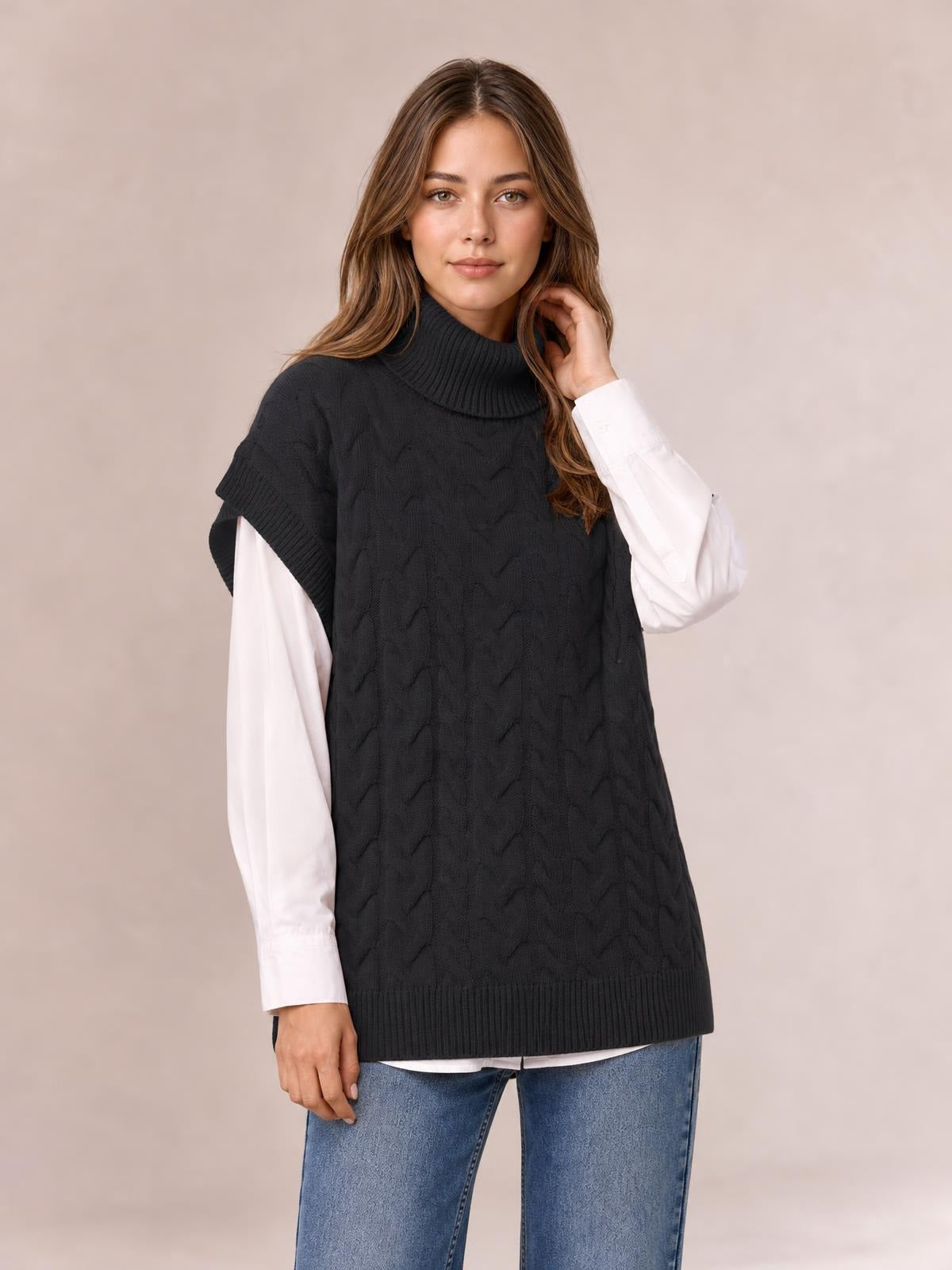 A model poses while touching her hair in a black chunky cable-knit turtleneck sweater with cap sleeves, ribbed trim, and a relaxed long silhouette.