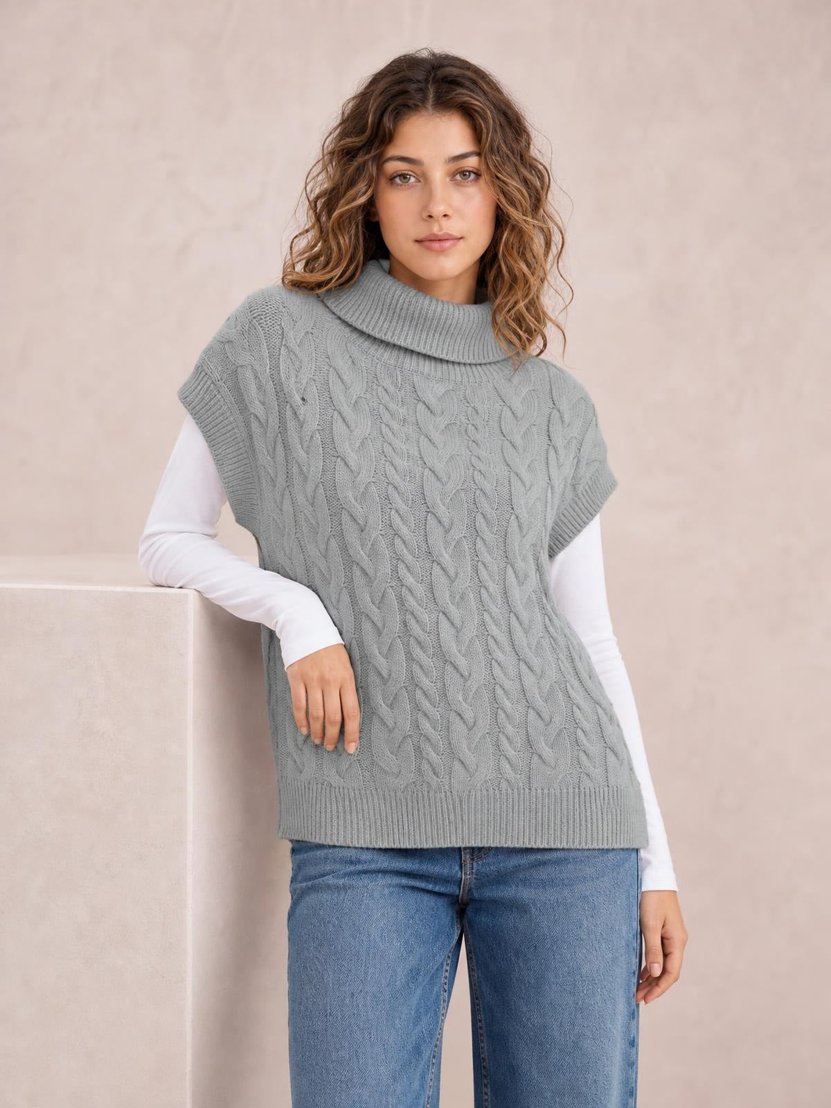 Model leans against a ledge wearing a gray chunky cable-knit turtleneck cap-sleeve sweater with ribbed trim and a relaxed, hip-length silhouette.