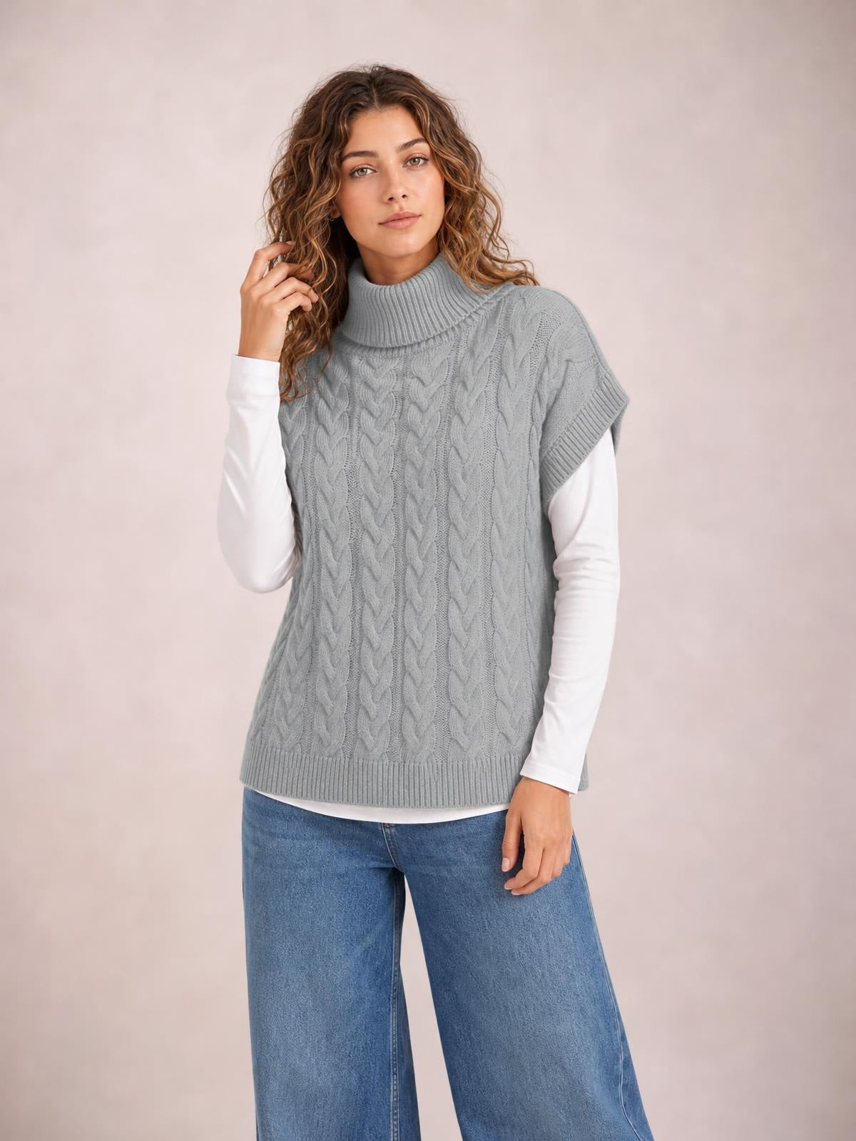 Model wears a gray chunky cable-knit turtleneck sweater with cap sleeves, ribbed trim, a straight relaxed silhouette, and layered white long sleeves.