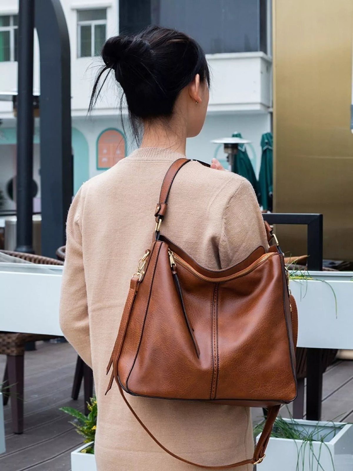 Model viewed from behind carries a medium classic brown PU leather tote with a slouchy silhouette, shoulder straps, zip top, and stitched panel construction.