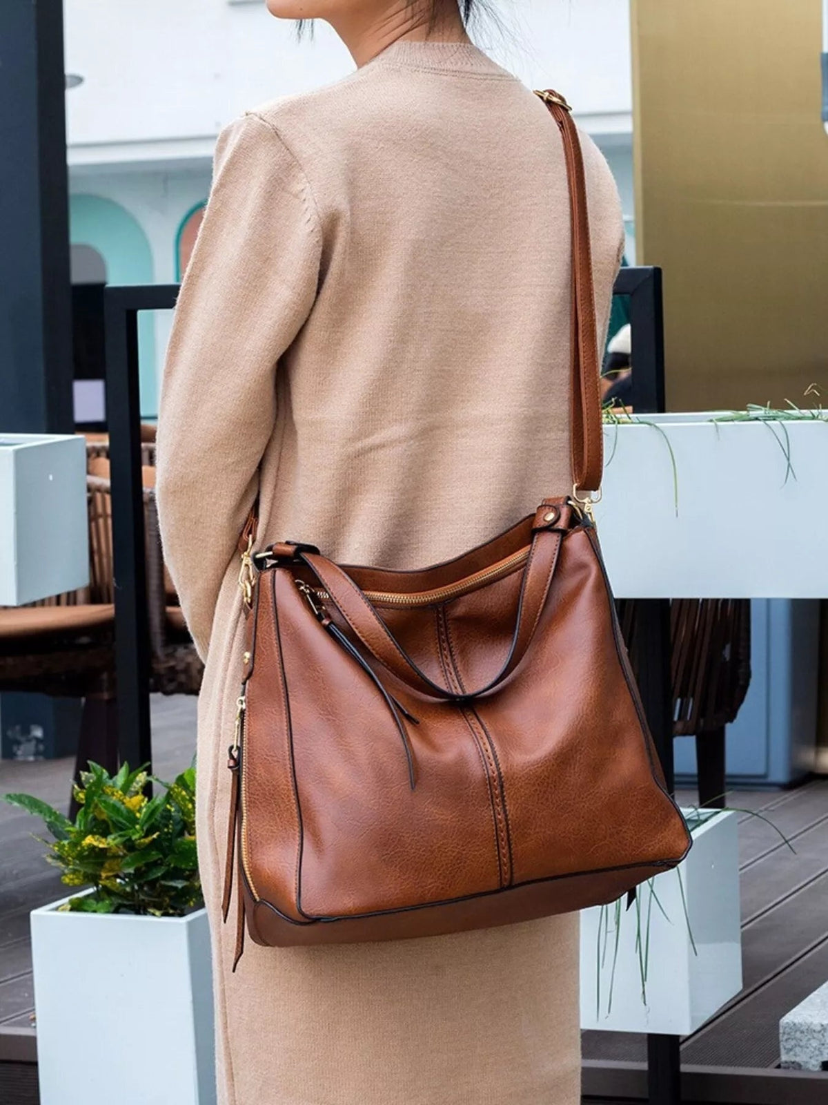 A model in a beige coat carries a medium brown PU leather tote bag with a structured silhouette, twin handles, long shoulder strap, and zippered top.