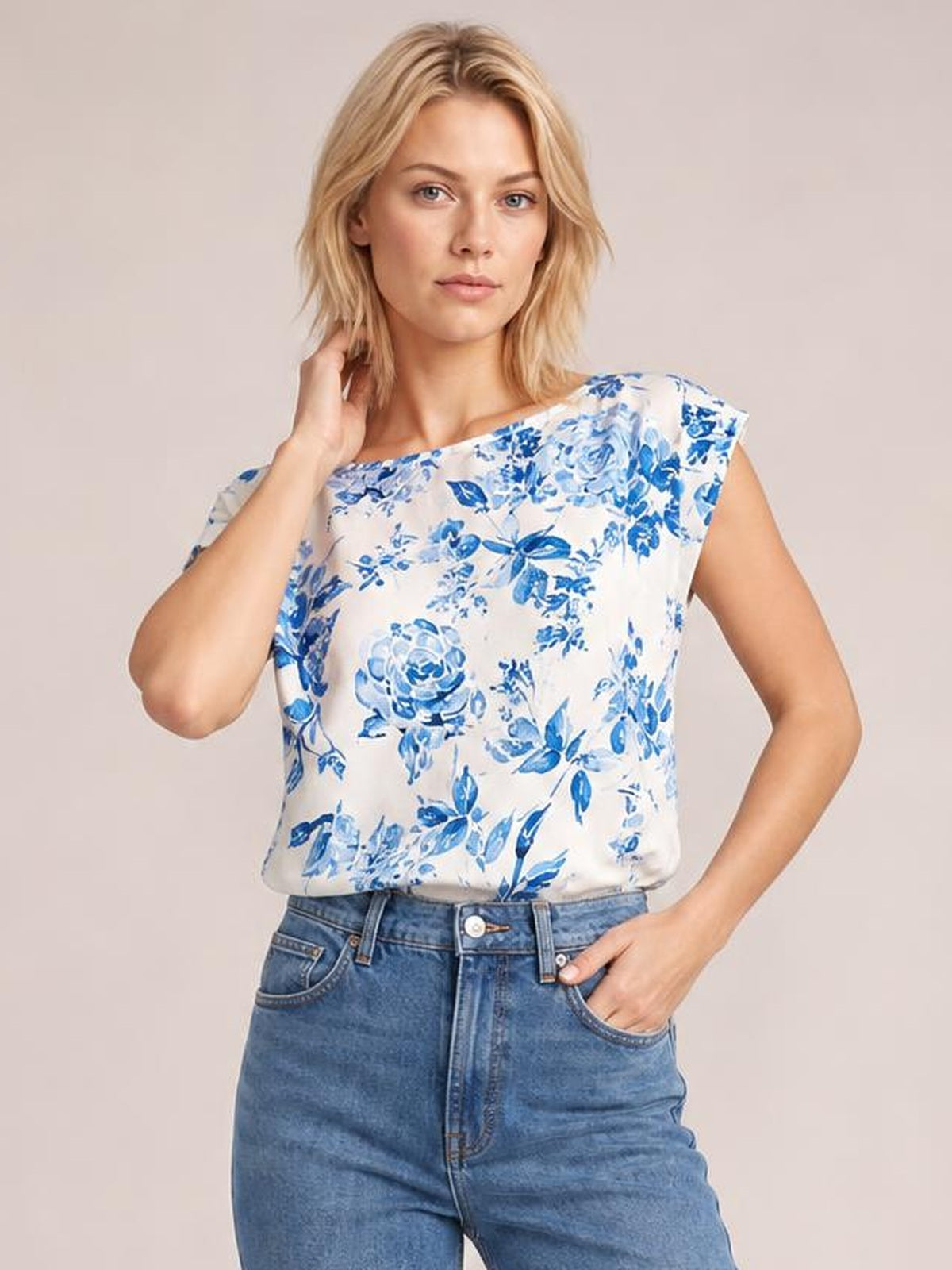 Model wearing a white and blue floral top with a wide neckline, short cap sleeves, and a relaxed straight silhouette, posed with one hand at her hair.
