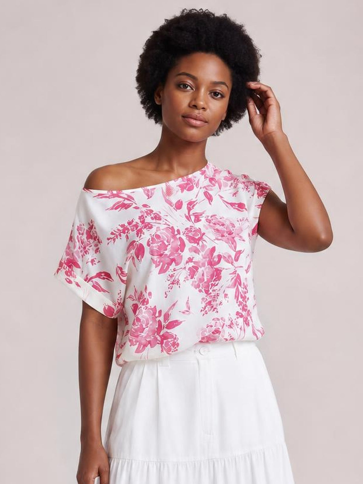 Model poses in a white and pink floral T-shirt with a wide neckline, relaxed silhouette, and short rolled sleeves, touching her hair.