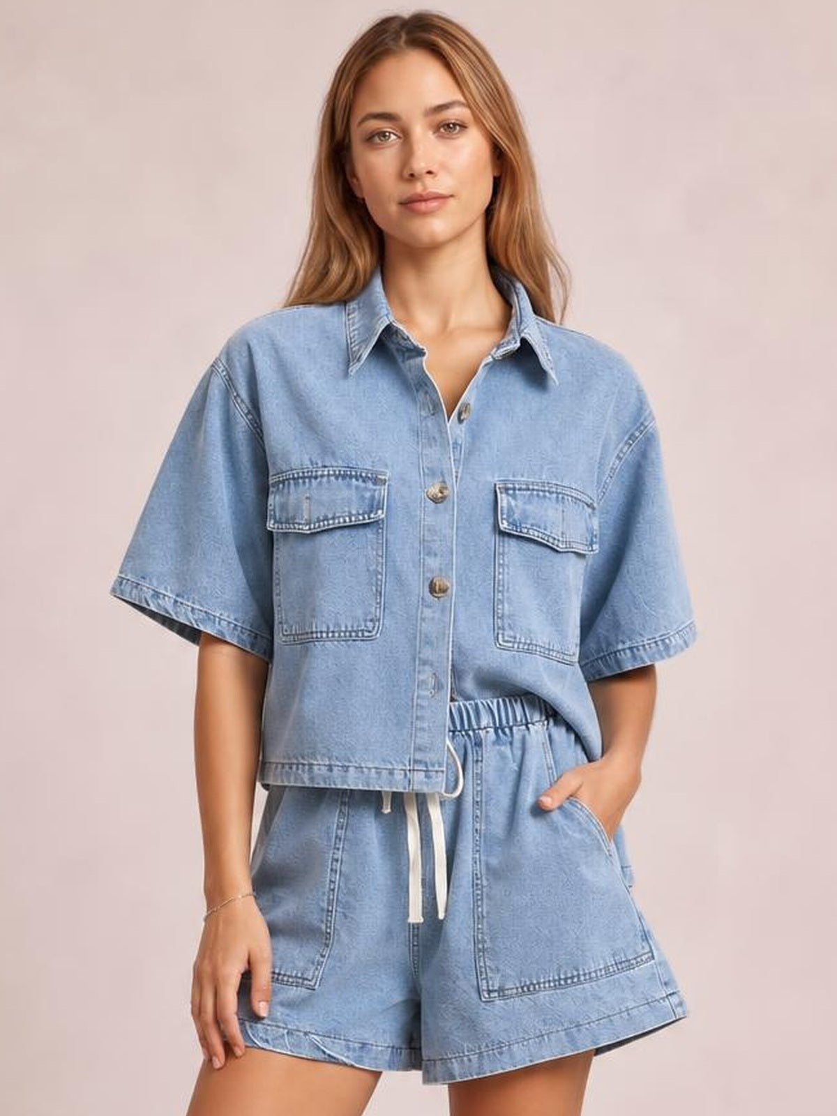 Model wearing a light blue denim set with a collared button-up top, flap chest pockets, short sleeves, and matching high-waisted drawstring shorts.
