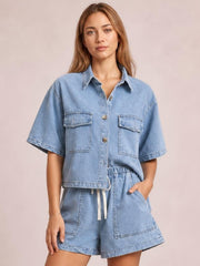 Model wearing a light blue denim set with a collared button-up top, flap chest pockets, short sleeves, and matching high-waisted drawstring shorts.